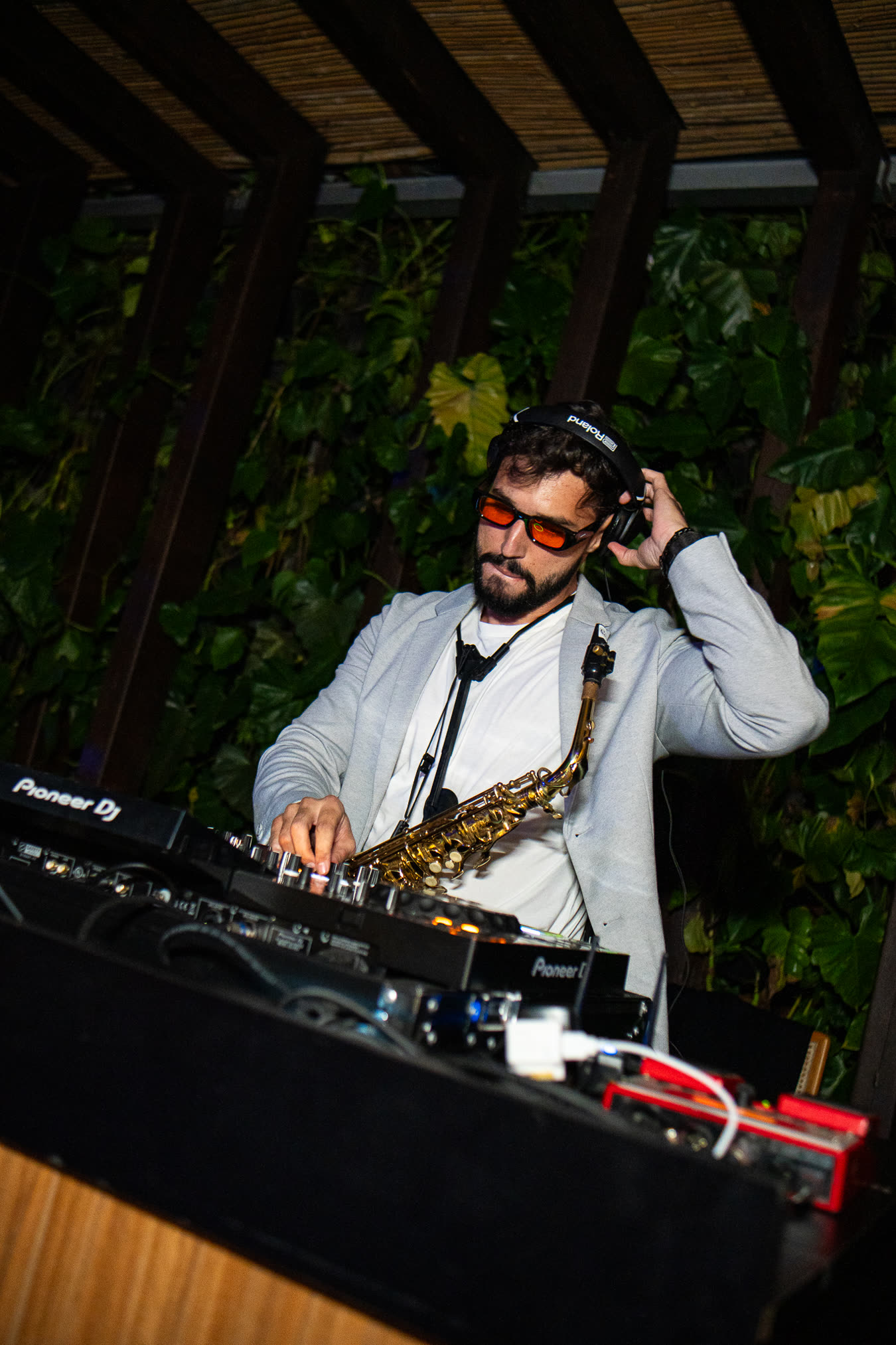 Bruno Daner de blazer claro atrás do mixer de DJ, com o saxofone pendurado